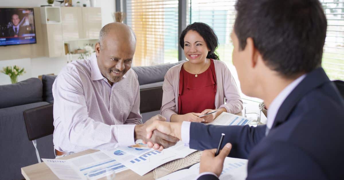 Two clients sit across from their banker at a desk in an office. One client is shaking the banker's hand while the other smiles.